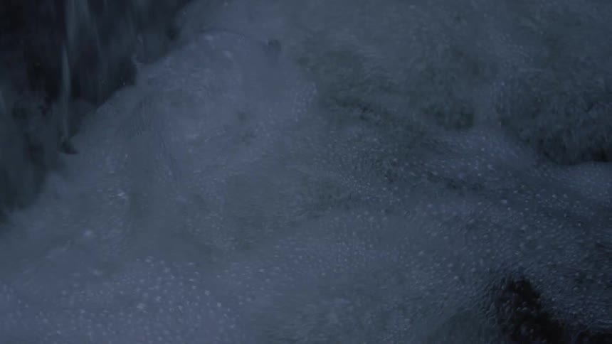 A moody close-up shot pans across a small waterfall as it powerfully cascades into churning, foamy water at night