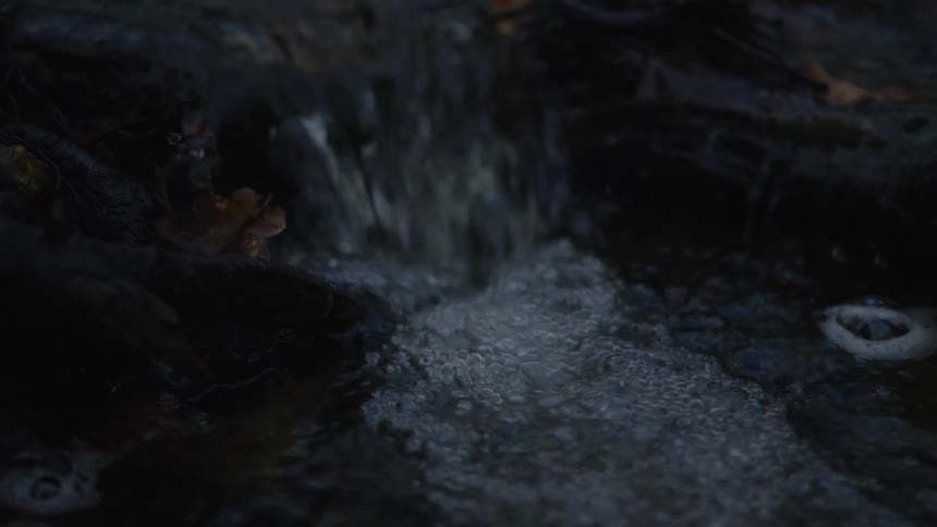 A close-up slow-motion shot of water bubbling and flowing down a dark, rocky stream at night