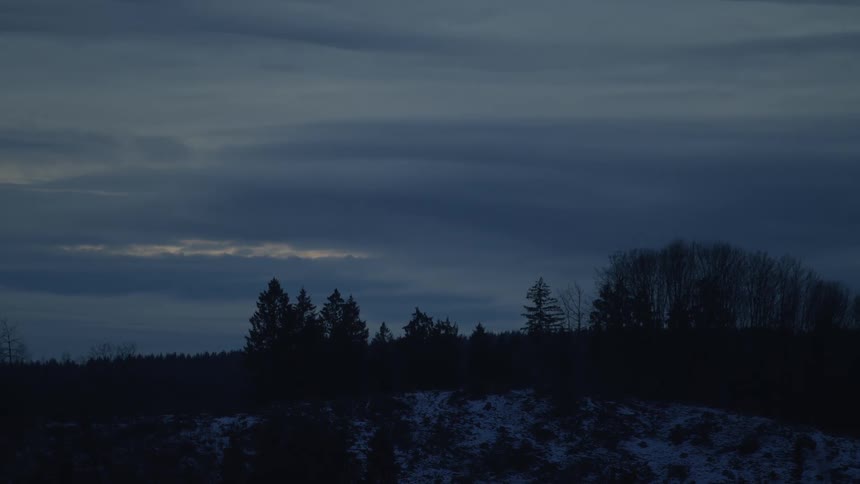 A somber, dark blue sky at dusk settles over a silhouetted treeline on a snowy winter evening