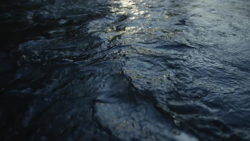 Dark, moody water ripples gently in slow motion with a glint of light on the surface