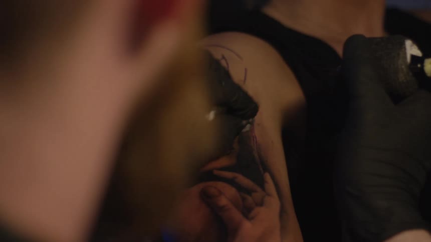 An over-the-shoulder view of a tattoo artist in black gloves meticulously inking a detailed portrait on a client's arm