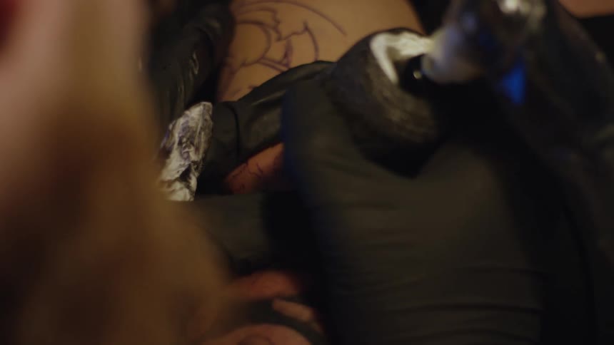 A tattoo artist in black gloves meticulously applies ink to a client's arm, outlining a portrait design in a dimly lit studio