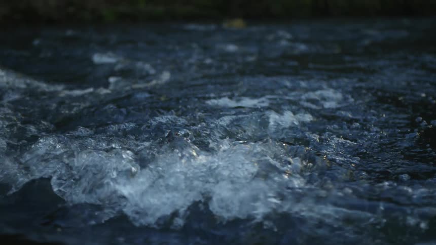 A close-up, slow-motion shot of dark river water churning and flowing in the low light of dusk