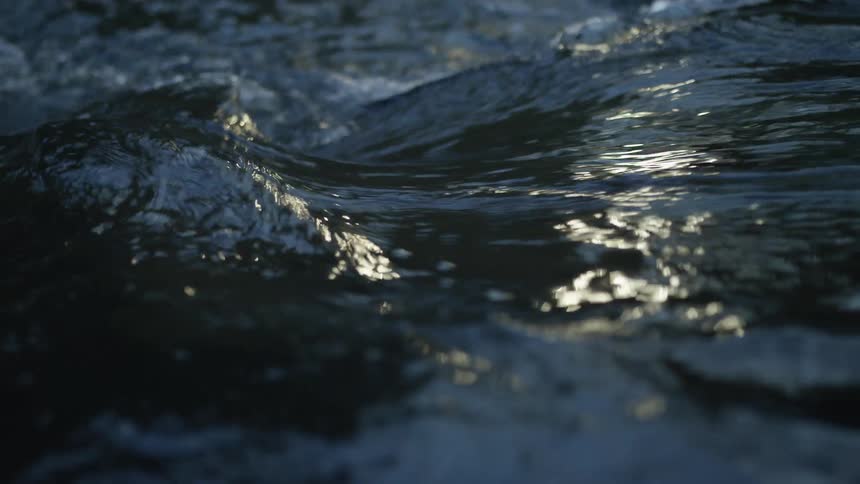 Slow motion close-up of dark, moody river water rippling and flowing with sunlight reflecting on the surface