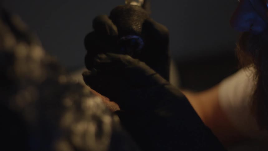 A close-up of a tattoo artist with a beard and glasses carefully applying ink to a client's skin in a dimly lit studio