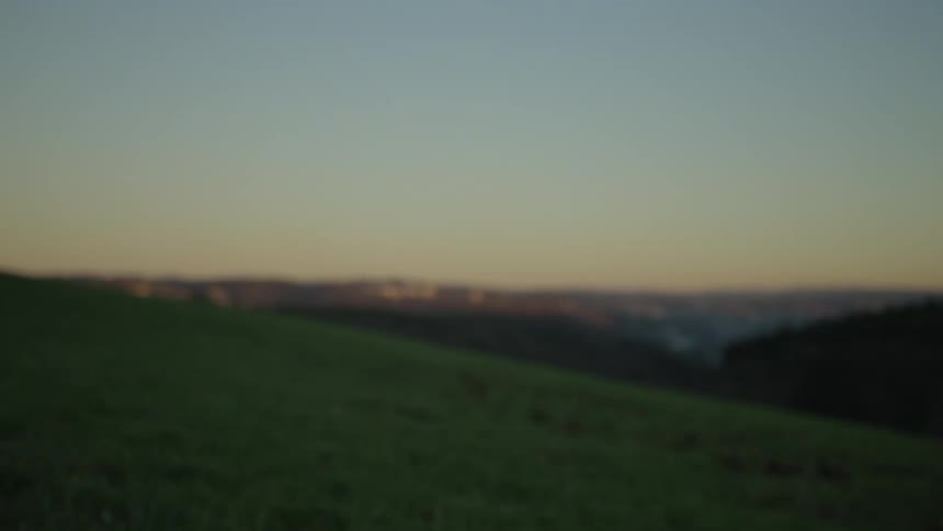 A slow tilt up from dark grass on a hillside reveals a blurry landscape and a clear sky at dusk