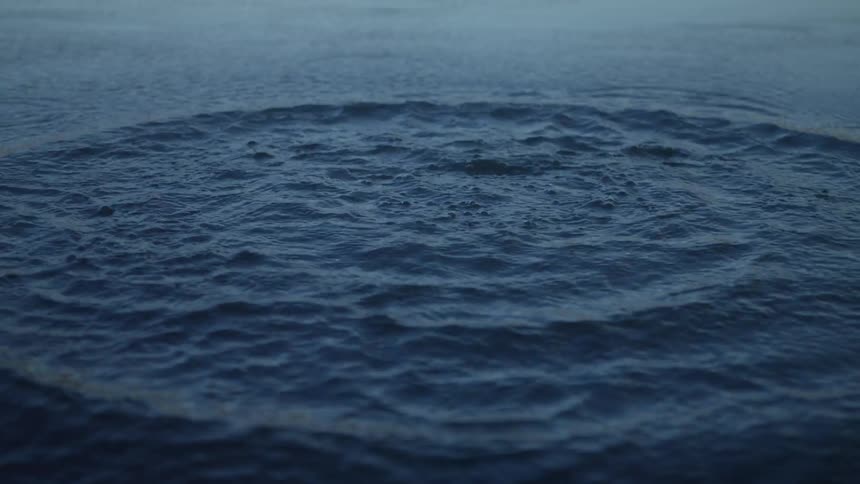 A stone is thrown into calm blue water, creating a dramatic splash and spreading ripples in super slow motion