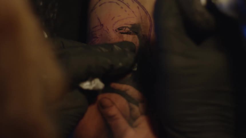 An overhead close-up shot shows a tattoo artist inking the details of a portrait onto a client's arm