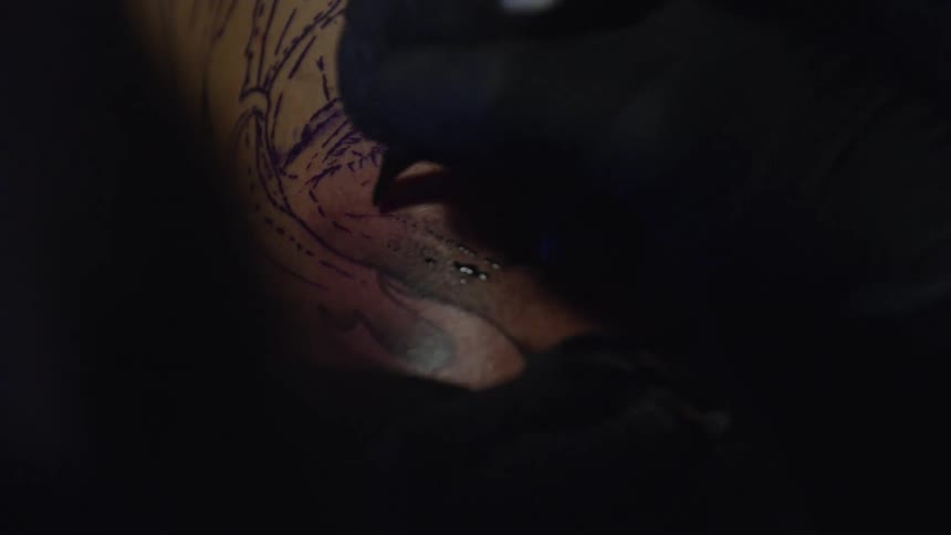 Close-up of a tattoo artist in black gloves meticulously inking a portrait onto a client's skin in a dark studio