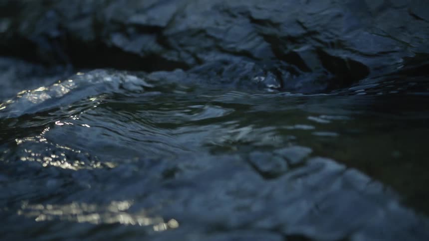 In extreme slow motion, clear water calmly flows over dark, smooth rocks, catching reflections of light
