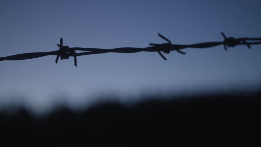 A slow pan across a silhouetted barbed wire fence against the dark evening sky