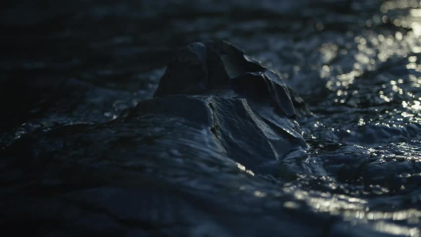 Dark, moody water flows in slow motion around a rock as light glistens off the surface