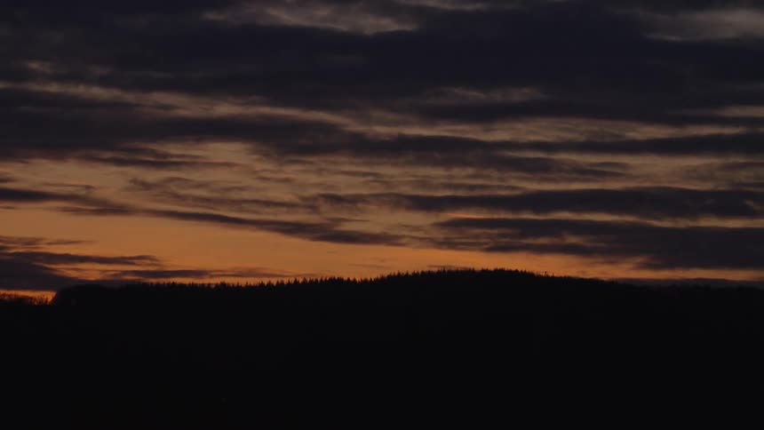 A moody timelapse of a dramatic orange sunset with dark clouds moving over a silhouetted forest treeline as night falls