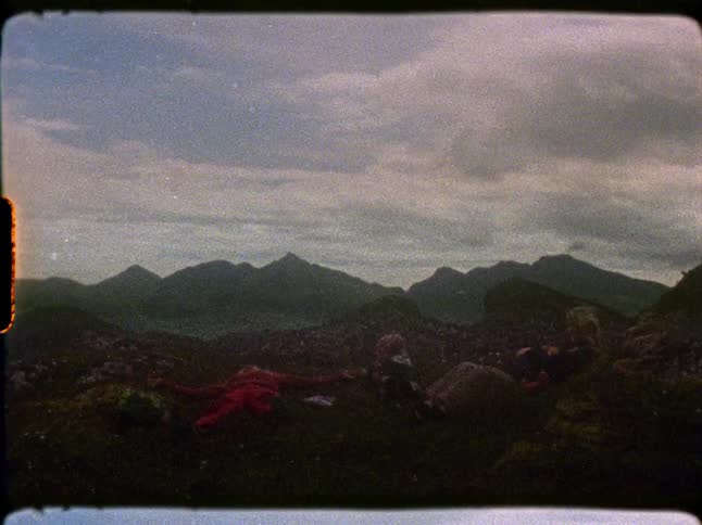 A vintage 8mm film captures young friends resting on a grassy mountaintop with a vast mountain range in the background
