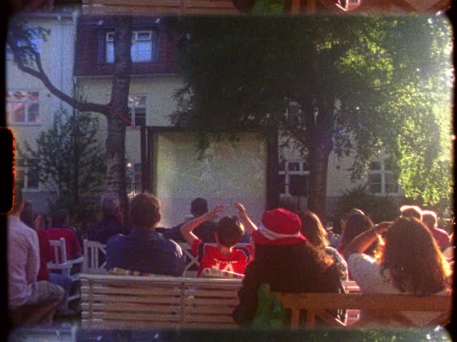 Nostalgic Super 8 footage shows a crowd gathered for an outdoor movie screening on a warm summer evening
