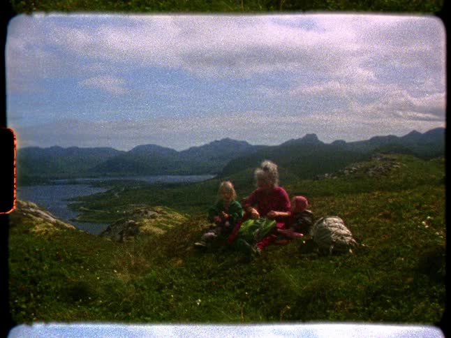 Vintage 8mm home movie footage shows a woman and two small children resting on a grassy hilltop overlooking a vast lake and mountains