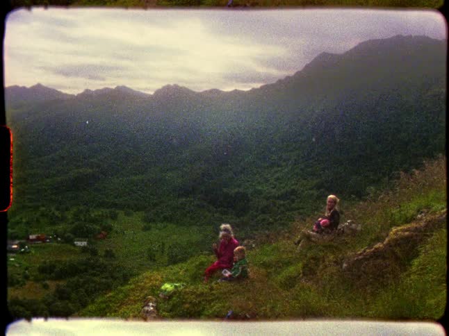 Vintage 8mm film of a mother and her children resting on a mountainside while hiking and enjoying the scenic valley view