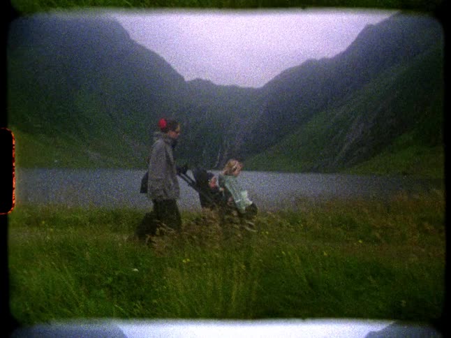 Vintage 8mm film captures a family walking with their child in a stroller along a path by a misty mountain lake