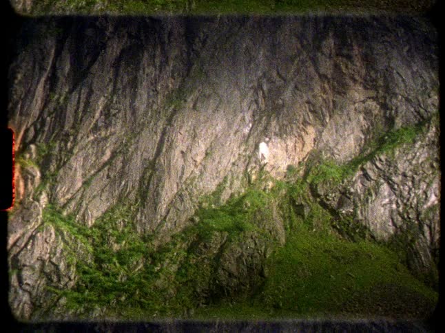 A close-up view of a waterfall cascading down a sheer rock face covered in green moss, captured on grainy vintage film