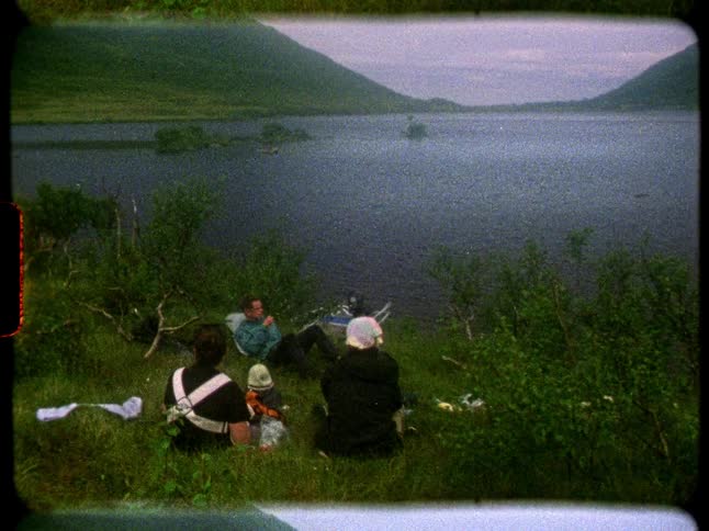 Vintage 8mm footage shows a family relaxing on a grassy hillside overlooking a serene lake and mountains