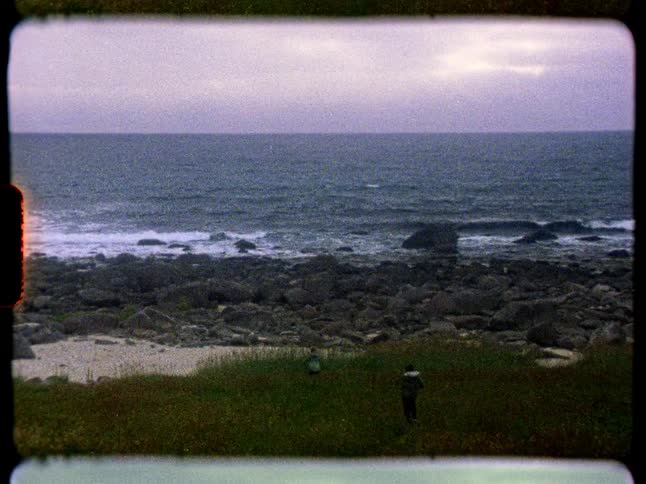 Grainy Super 8mm film shows two children walking through a grassy field towards a rocky ocean beach