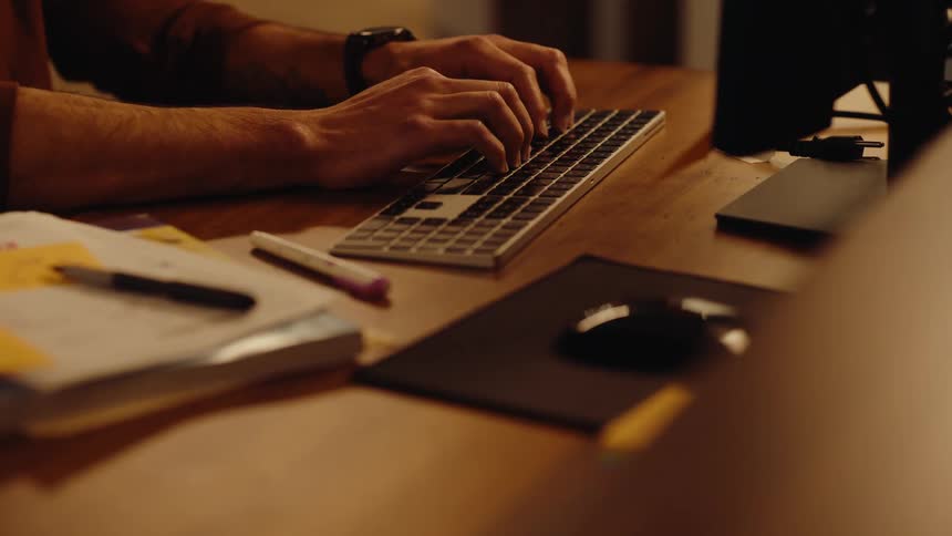 Close-up of a man's hands typing on a modern keyboard while working late at his wooden desk
