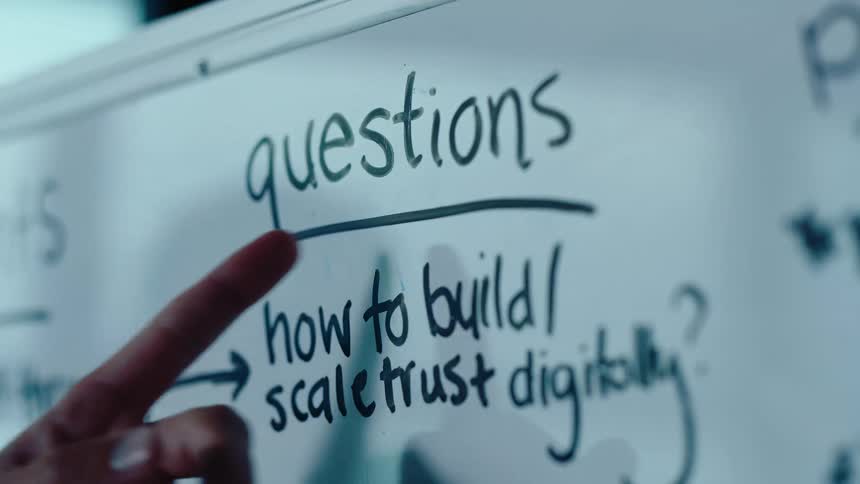 A presenter's hand points to strategic questions about building digital trust written on a whiteboard during a business meeting