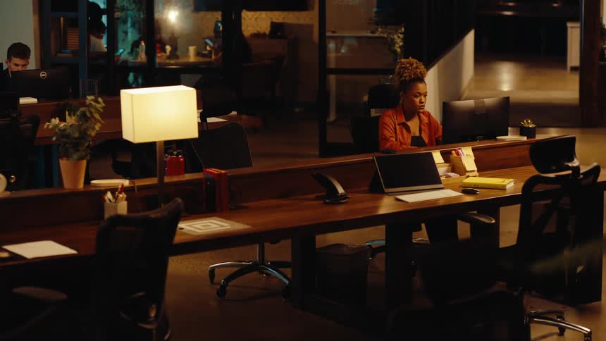 A woman works diligently at her computer in a dimly lit, modern open-plan office late at night
