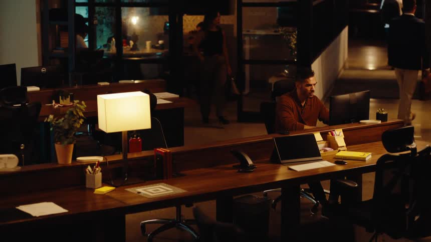 A man works late at his desk in a modern, dimly lit office while his colleagues leave for the night