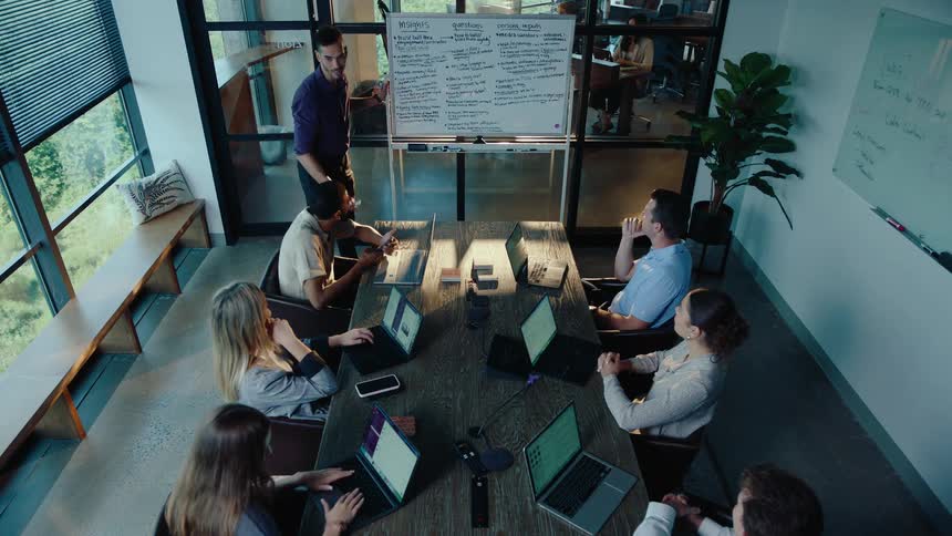 A diverse team of business professionals collaborates in a modern conference room as one man leads a presentation on a whiteboard