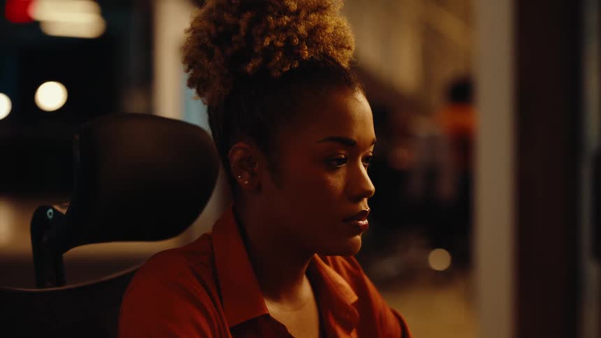 A close-up side profile of a focused young Black woman working late at night in a modern office