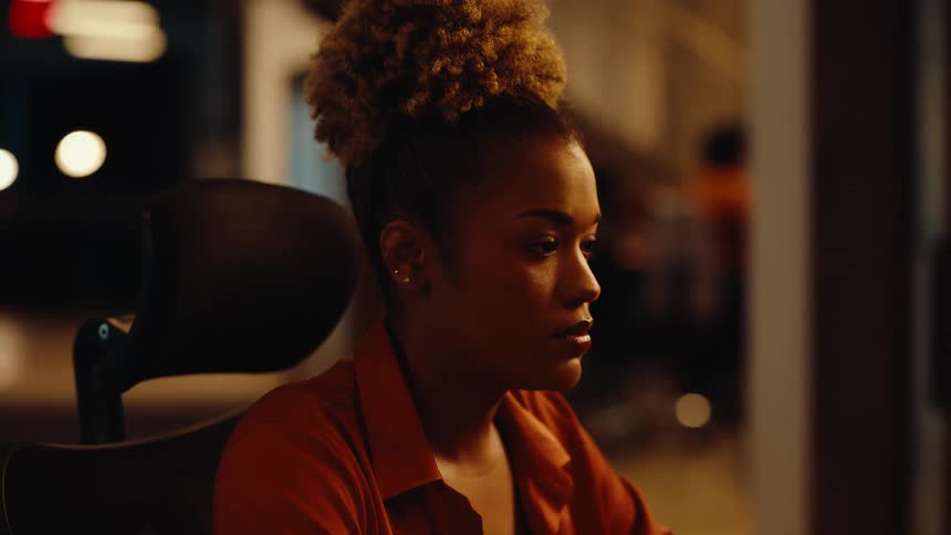 A focused young woman with a high bun hairstyle concentrates on her work late at night in a dimly lit office