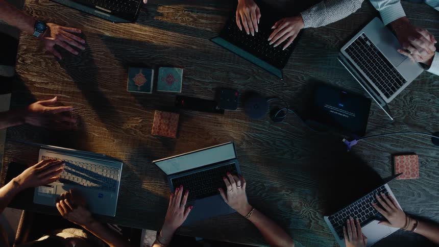 Slow zoom out from a top-down view of a diverse group of colleagues collaborating and typing on their laptops around a wooden conference table