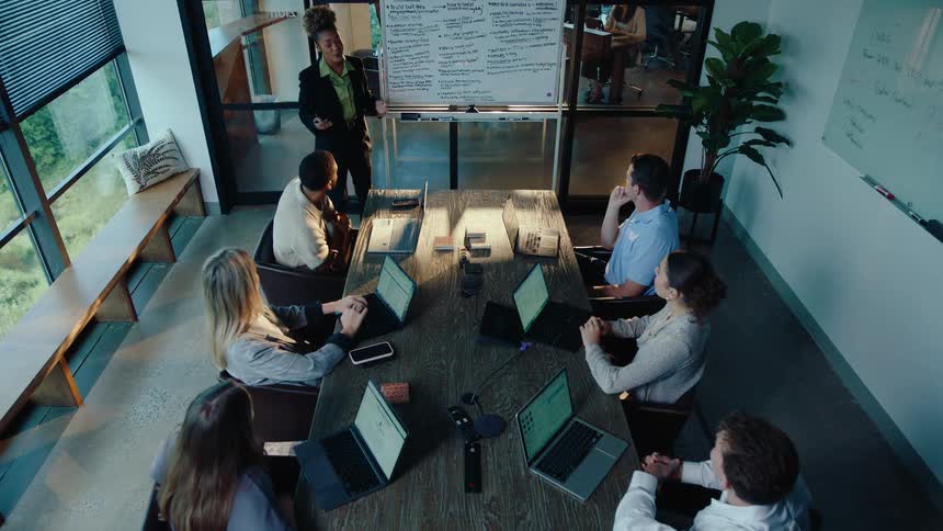 A businesswoman leads a diverse team in a brainstorming session, presenting ideas on a whiteboard in a modern conference room