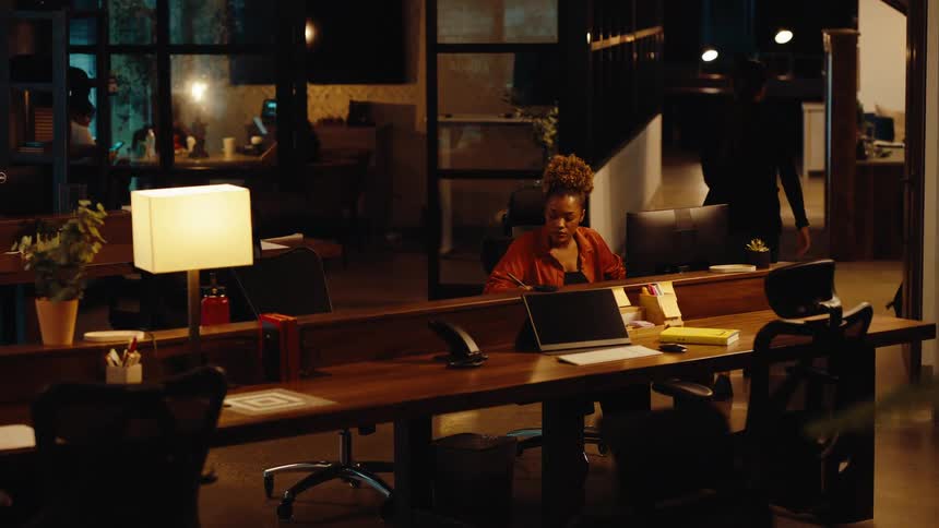 A woman leaves the office for the evening, waving goodbye to her colleague who continues to work late in a modern, dimly lit workspace