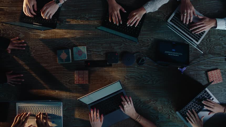 A top-down, slow zoom-out reveals a diverse team of professionals diligently typing on their laptops during a collaborative meeting around a wooden table