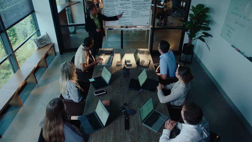 High-angle view of a woman leading a business meeting, presenting project details from a whiteboard to her colleagues in a modern office
