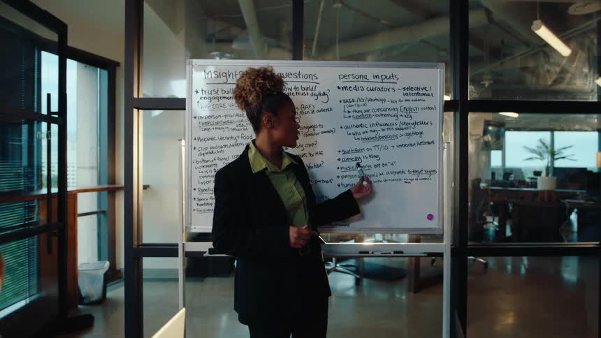A Black woman leads a business presentation on a whiteboard to her diverse team in a modern conference room as they take notes on their laptops