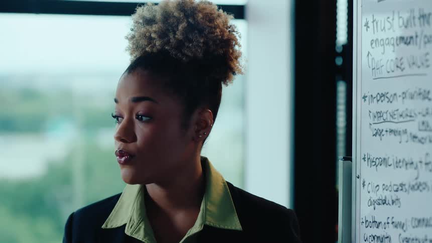 A professional woman with her hair in a bun gives a presentation and points to notes on a whiteboard in an office