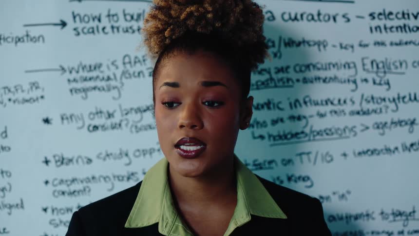 A professional Black woman explains a business strategy written on a whiteboard in a meeting