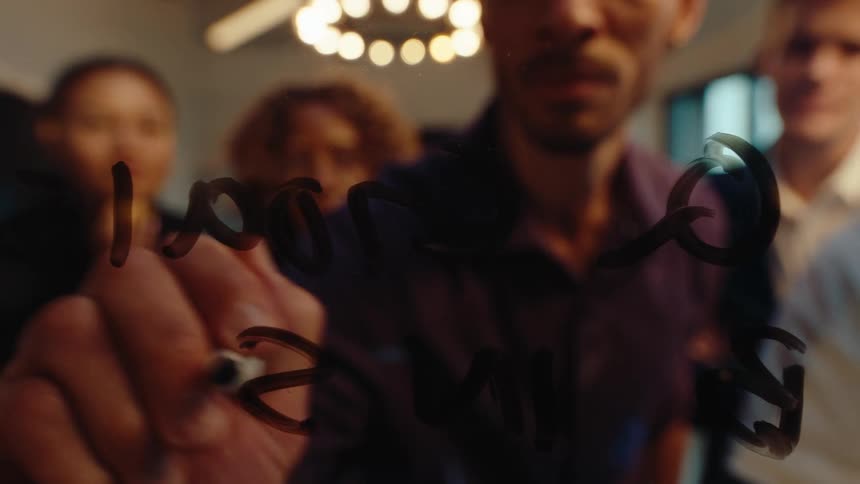 Close-up of a businessman's hand writing positive company results on a glass board while colleagues watch in the background
