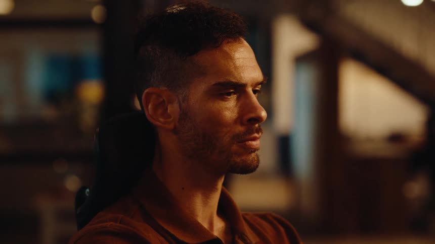 Close-up profile of a pensive man concentrating intently in a dimly lit, modern office space at night