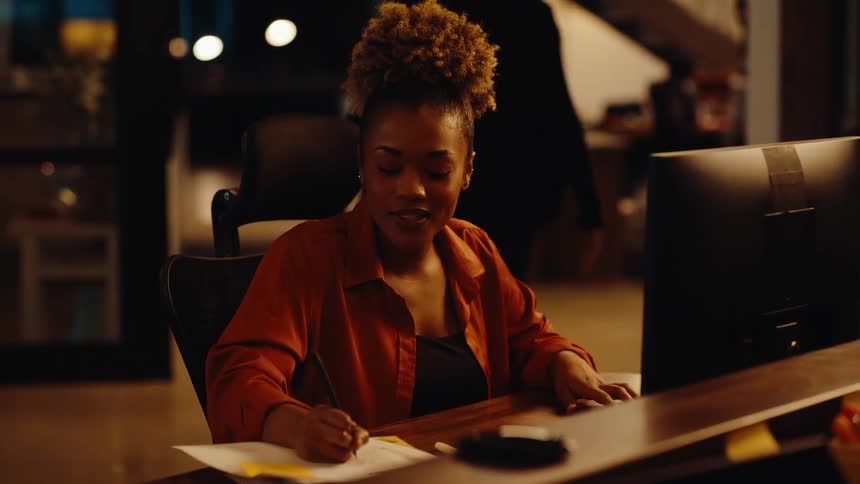 A focused young businesswoman works late at her desk in a modern, dimly lit office, typing and taking notes while a colleague walks by in the background