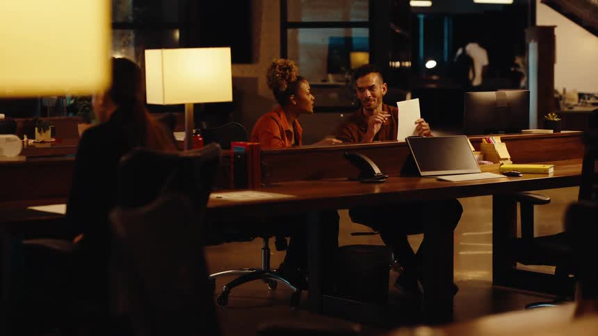 Diverse colleagues smile and collaborate on a project while reviewing documents in a stylish, modern office late at night
