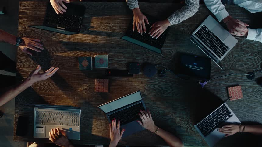 A top-down view shows a team of professionals collaborating and typing on laptops during a meeting around a wooden table