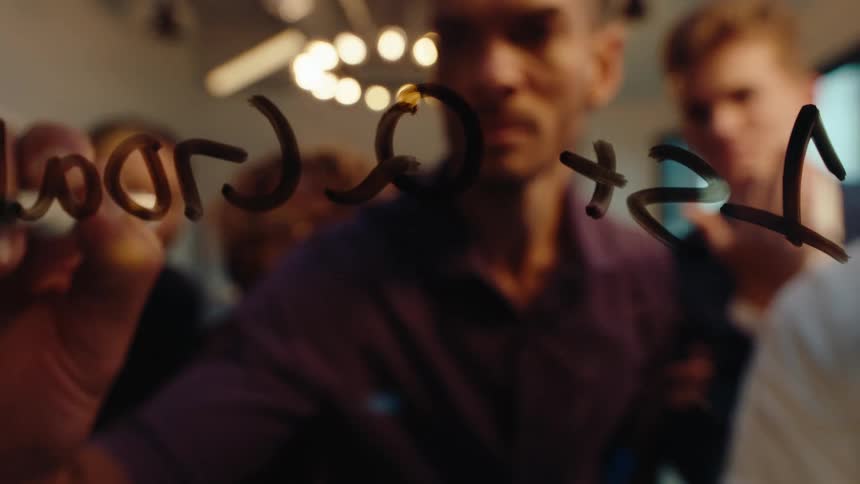 Close-up slow motion shot of a man's hand writing '4th Quarter Forecast' on a glass board as his team watches in the background