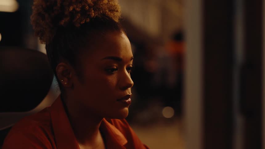 A young businesswoman with an afro puff bun works diligently at her computer late at night in her office