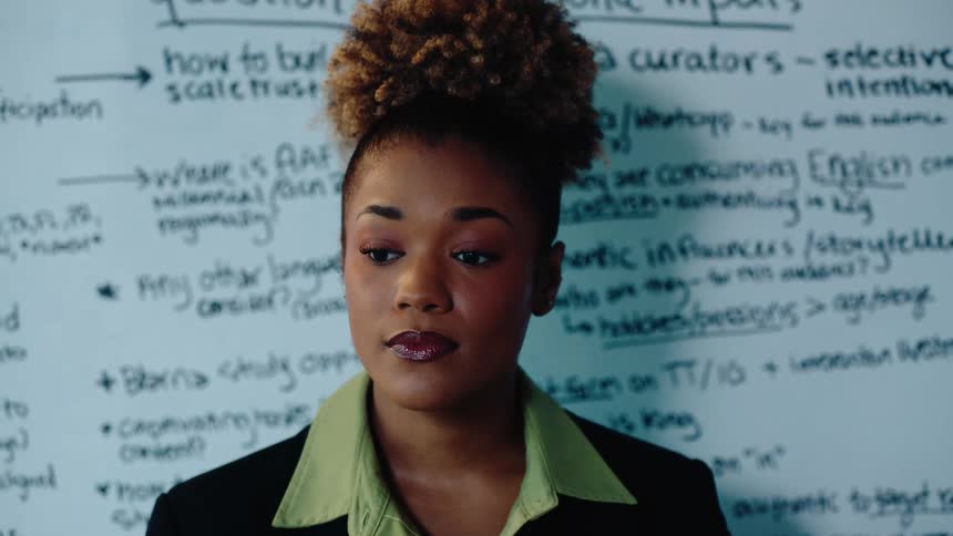 An articulate Black woman with curly hair points to a whiteboard while giving a presentation in a corporate setting