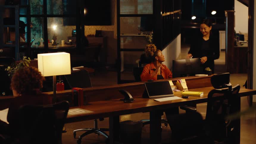 A diverse group of colleagues work late in a modern office, one woman stops to say goodbye to her coworker before leaving for the night