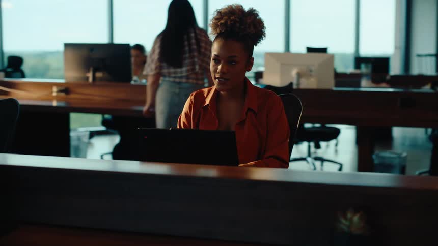 A young African American businesswoman diligently works on her laptop in a dynamic and modern open-plan office